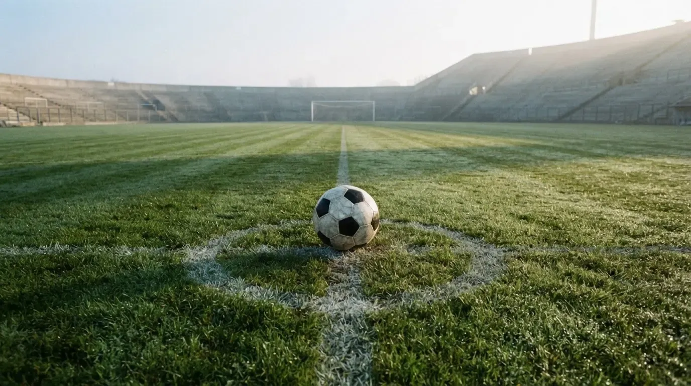 Ein Fussball liegt auf dem Anstosspunkt eines leeren Rasenplatzes im frühen Morgenlicht, dahinter erhebt sich eine menschenleere Tribüne.