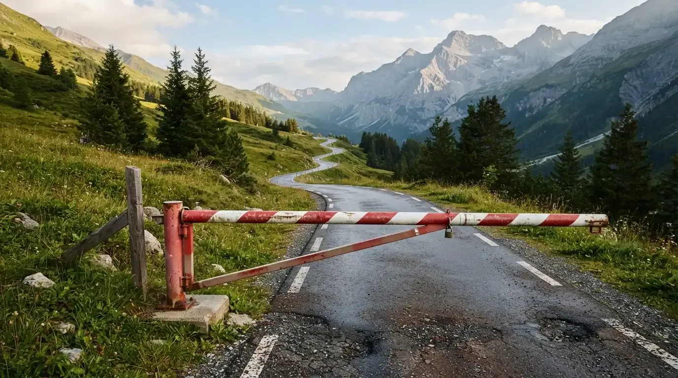 Ein Schlagbaum sperrt eine Landstrasse in den Bergen, dahinter windet sich der Weg zwischen grünen Hängen.