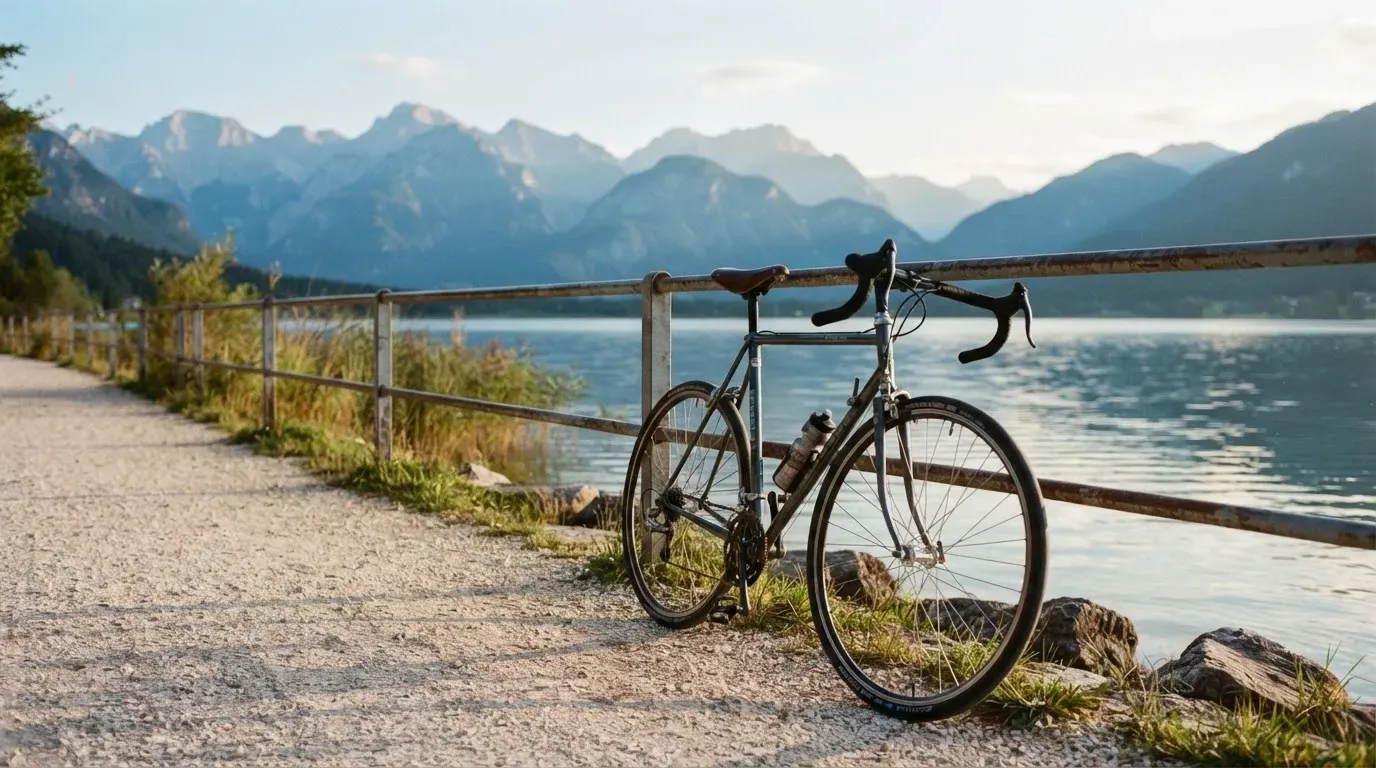 Ein Rennrad lehnt an einem Geländer am Seeufer, dahinter liegt ruhiges Wasser vor einer Bergkette.