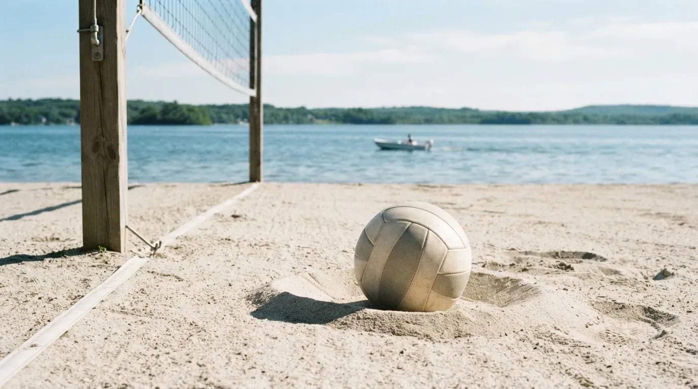 Ein Volleyball liegt im Sand eines Spielfelds am Seeufer, im Hintergrund gleitet ein Boot über das Wasser.