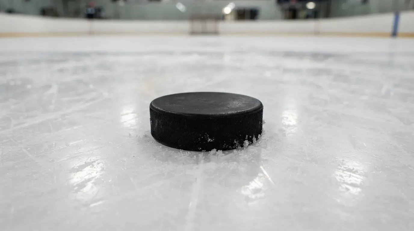Ein Eishockeypuck liegt in Grossaufnahme auf der Eisfläche, feine Kratzer im Eis glänzen im hellen Hallenlicht.
