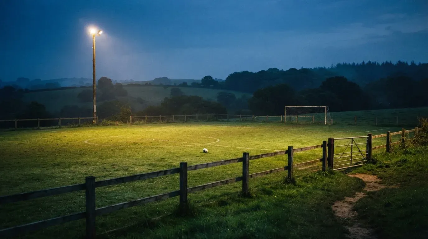 Ein kleiner Dorf-Fussballplatz liegt in der Dämmerung unter einem hohen Flutlichtmast, am Spielfeldrand steht ein Holzzaun.