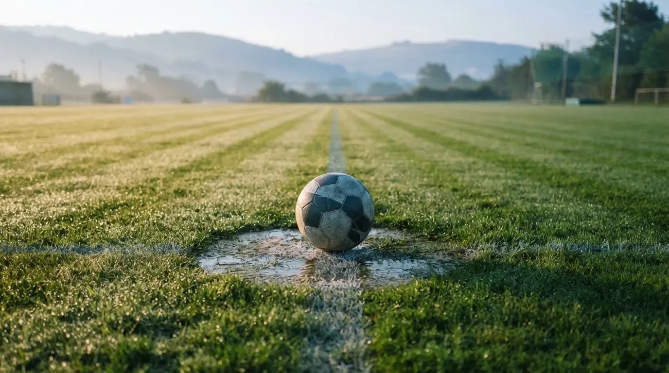 Ein Fussball liegt am frühen Morgen auf dem Anstosspunkt eines taunassen Rasens, im Hintergrund erheben sich Hügel im Dunst.