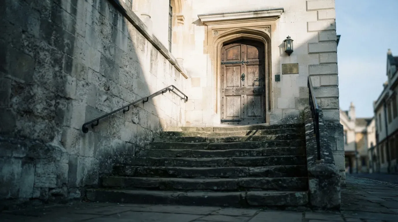 Eine Steintreppe führt zu einer schweren Eichentür in einer hellen Fassade, die Stufen liegen im Morgenschatten.