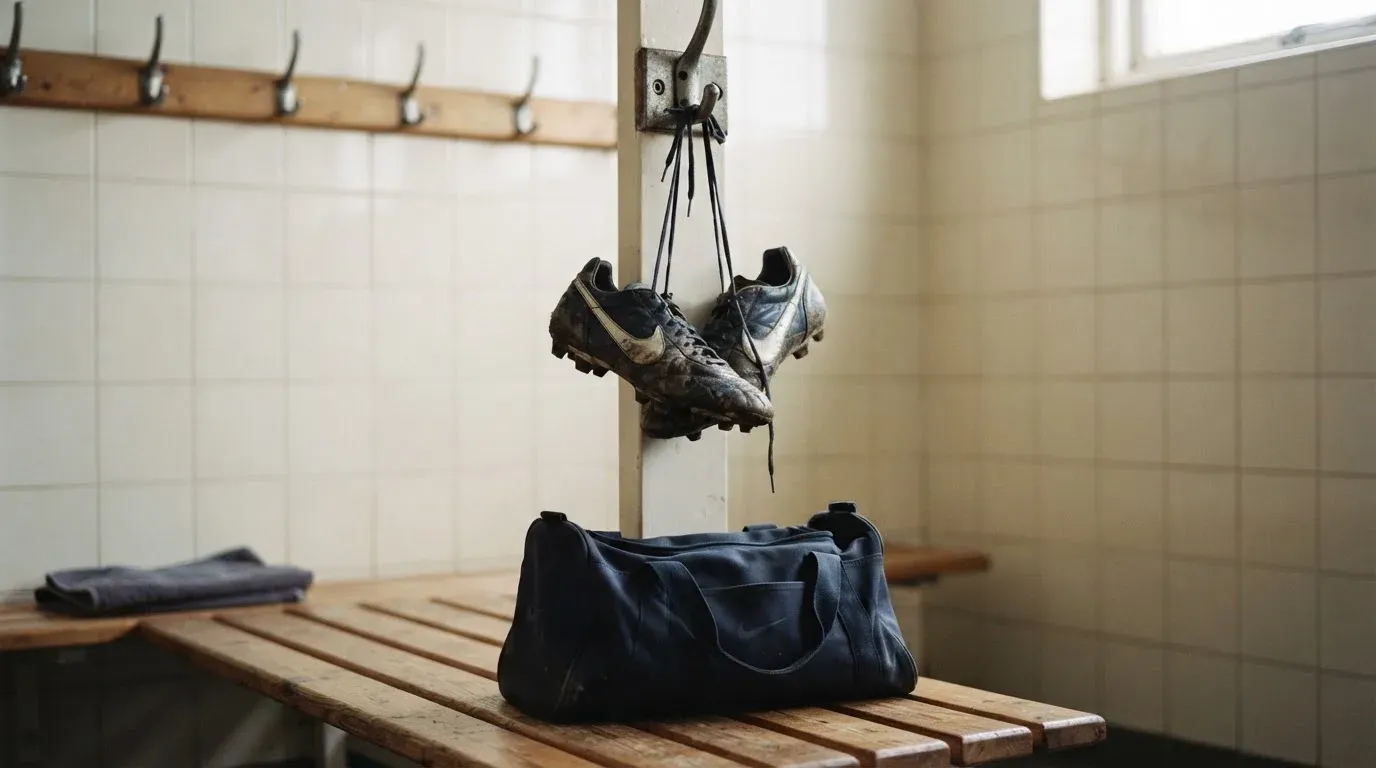 Ein Paar Fussballschuhe hängt an einem Haken in einer ruhigen Umkleide, darunter steht eine Sporttasche auf einer Holzbank.