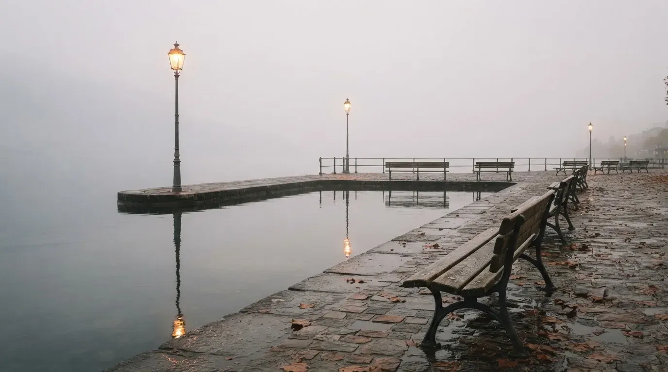 Eine Seepromenade mit leeren Bänken an einem nebligen Morgen, im Wasser spiegeln sich die Lichter der Uferlaternen.
