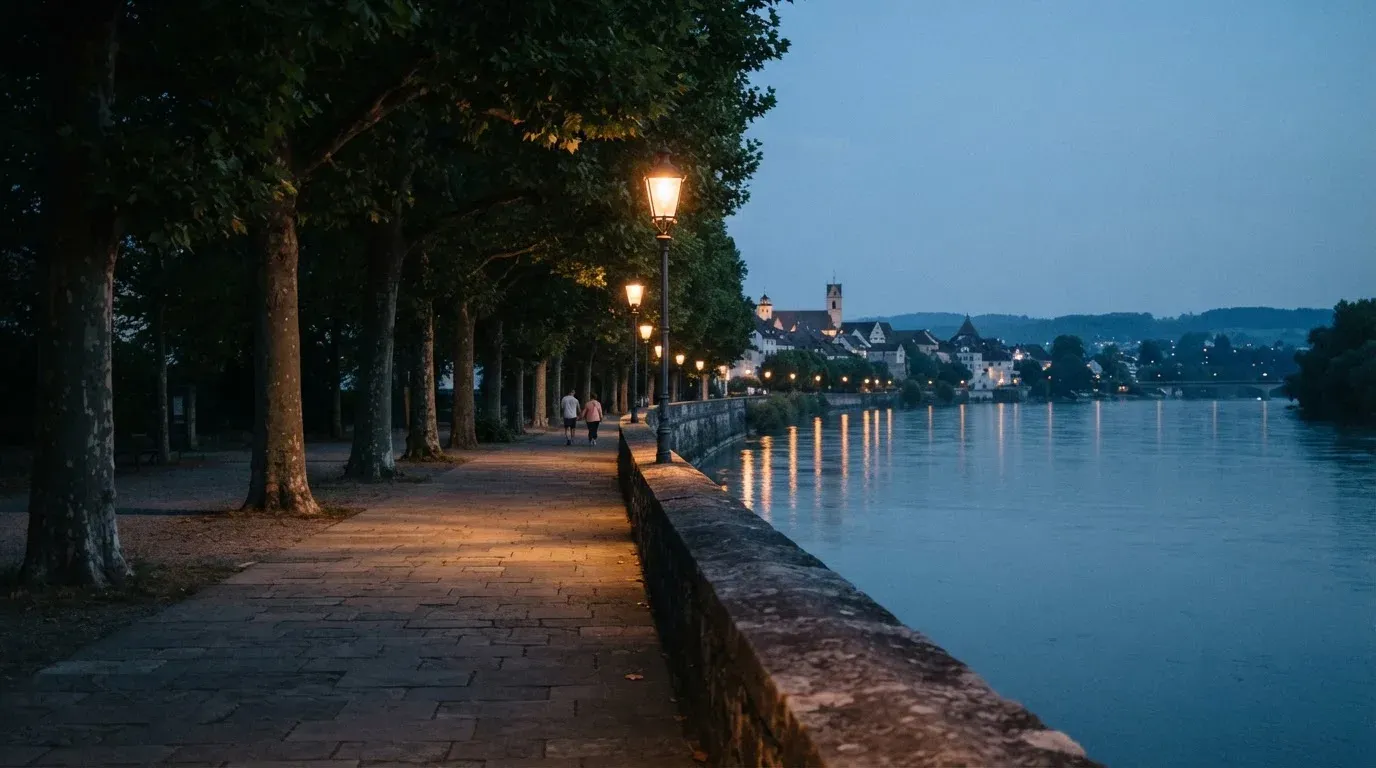 Eine Rheinpromenade mit hohen Bäumen am Abend, das Wasser spiegelt das Licht der ersten Laternen.