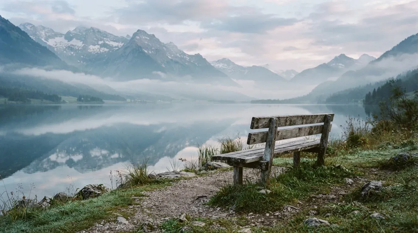 Eine Holzbank an einem Seeufer mit Blick auf eine Bergkette im Morgendunst, das Wasser ist spiegelglatt.