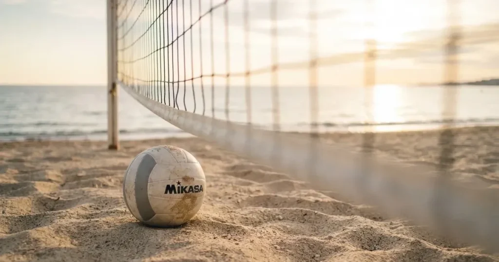 Ein Volleyball liegt auf feinem hellem Sand, ein gespanntes Netz verschwimmt dahinter, das Licht kommt von einem weiten Meer.