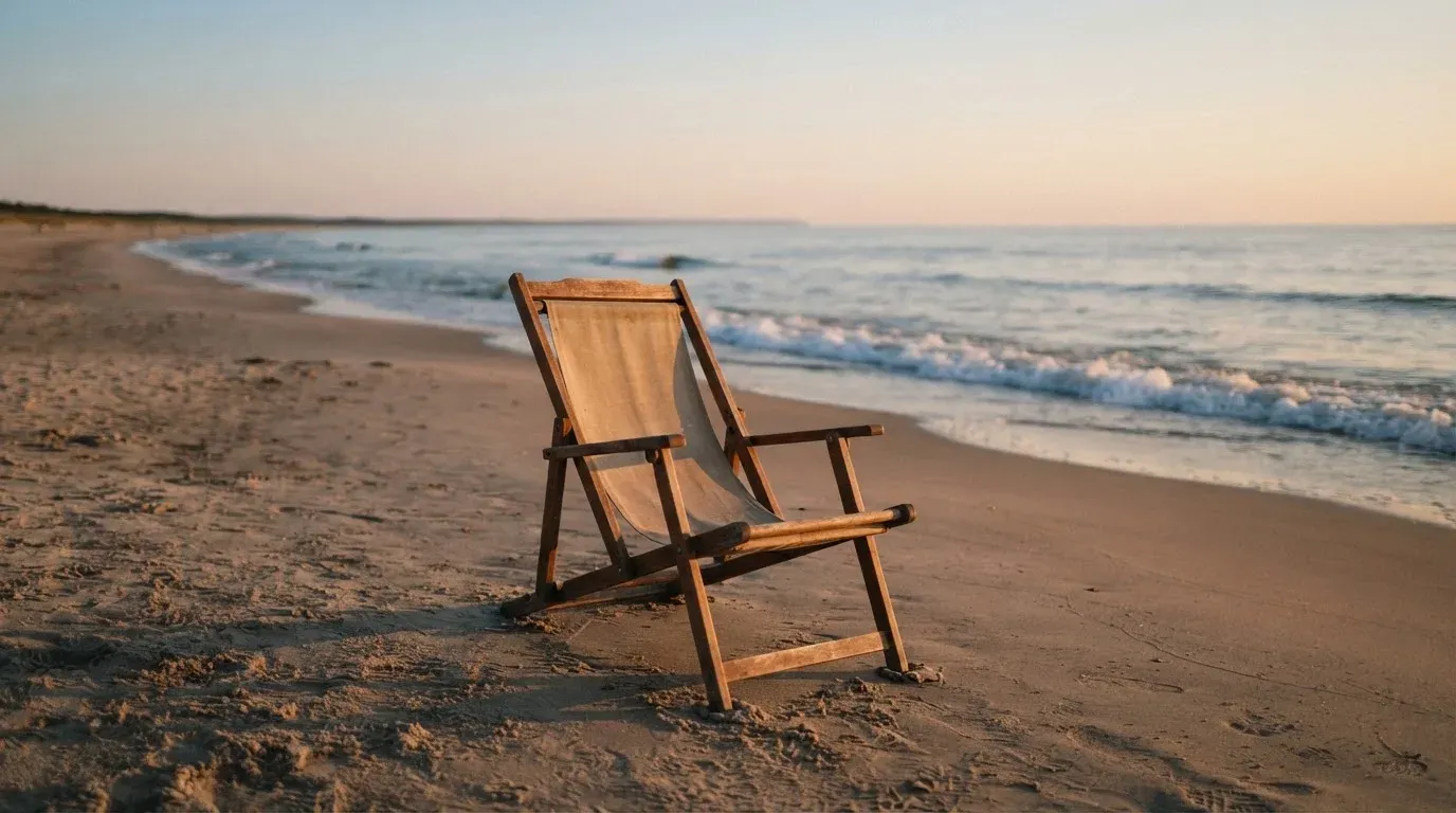 Ein leerer Liegestuhl aus Holz steht am Rand eines Sandstrands, Abendlicht und ruhige Wellen im Hintergrund.
