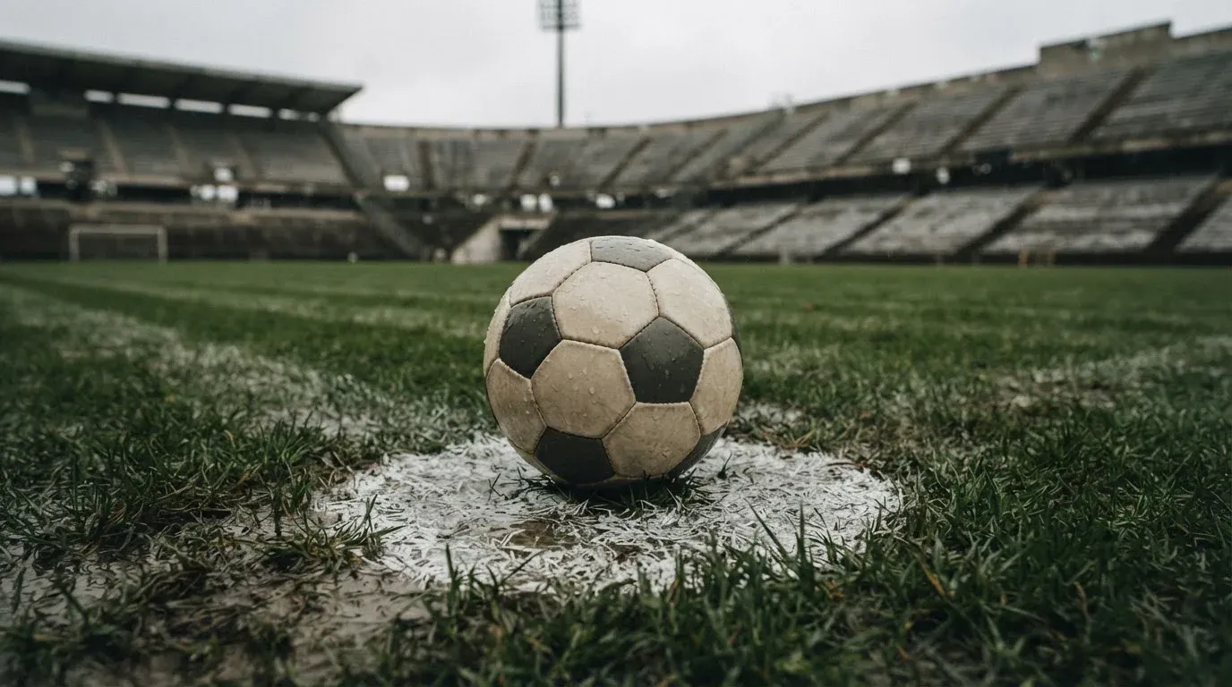 Ein Fussball liegt auf dem Anstosspunkt eines nassen Rasens, dahinter eine leere Tribüne unter grauem Himmel.