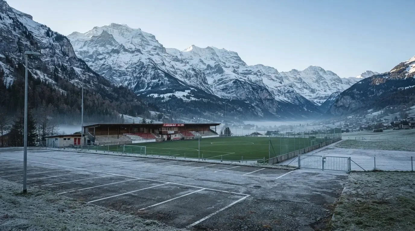 Schneebedeckte Berge ragen hinter einem kleinen Stadion in einem Alpental auf, im Vordergrund ein leerer Parkplatz am Morgen.