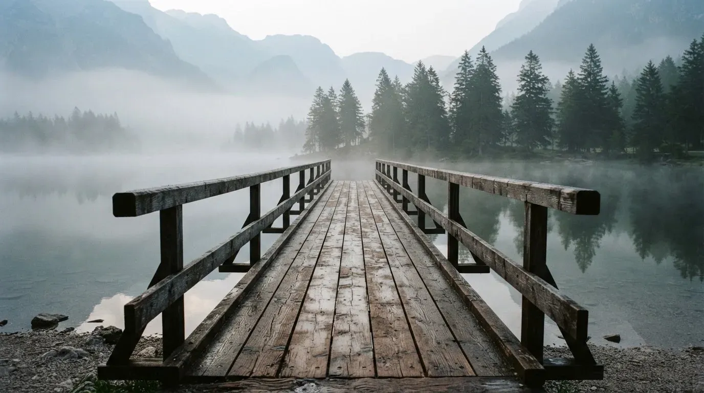 Ein Holzsteg führt über einen ruhigen Bergsee im Nebel, am anderen Ufer stehen dunkle Tannen.