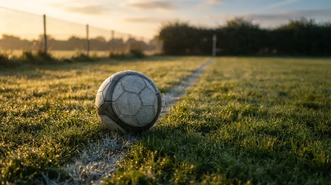 Ein Fussball liegt im Tau auf der Mittellinie eines Dorfplatzes bei Sonnenaufgang, der Rasen glitzert.