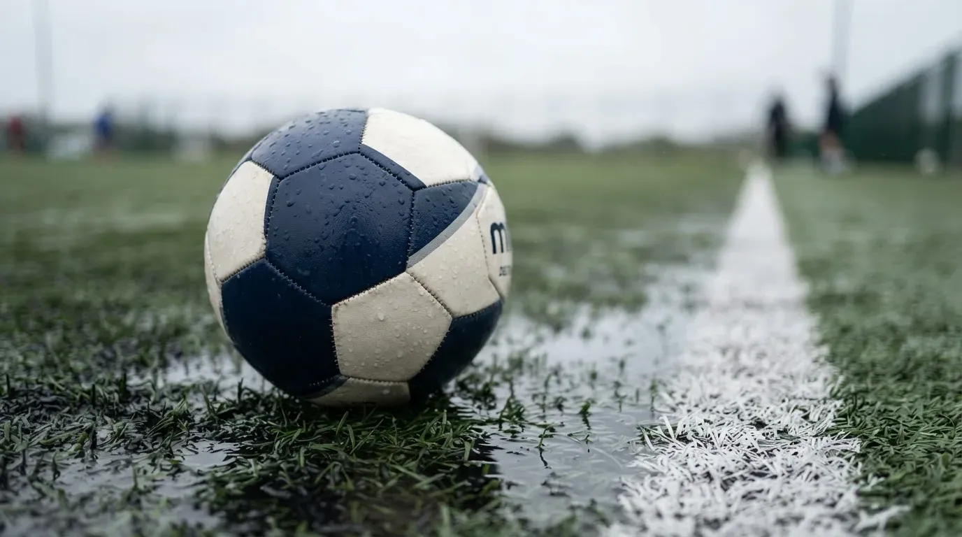 Ein Fussball liegt auf nassem Kunstrasen neben einer weissen Linie, Pfützen spiegeln den Himmel.