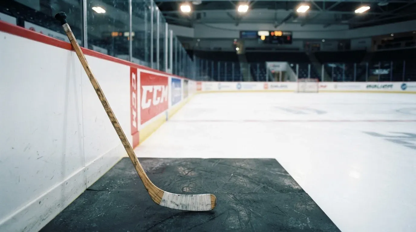 Ein Eishockeyschläger lehnt an der Bande, in der Ferne liegt die hell beleuchtete Eisfläche einer Halle unter Flutlicht.