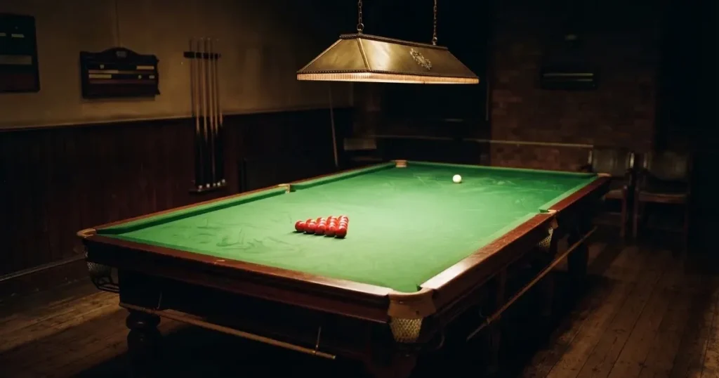 Ein Snookertisch mit grünem Tuch und einem Dreieck aus roten Kugeln unter einer tief hängenden Lampe in einer leeren Halle.