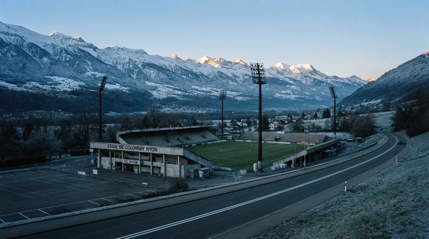 Schneebedeckte Berge erheben sich hinter einem Fussballstadion in einem Schweizer Tal, davor liegt eine leere Strasse im Morgenlicht.