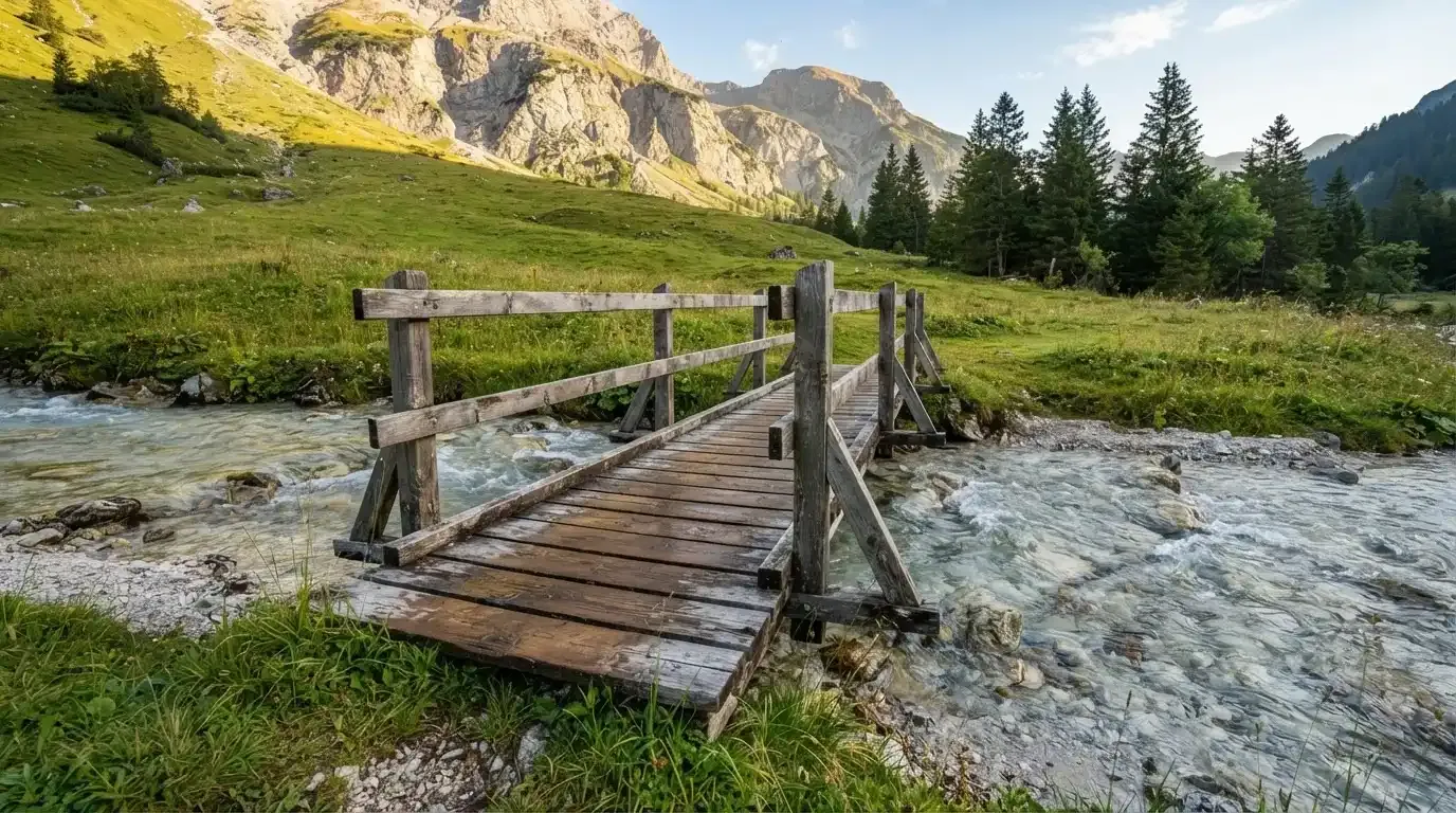 Ein Holzsteg führt über einen klaren Bergbach, im Hintergrund Wiesen und steile Hänge im Morgenlicht.