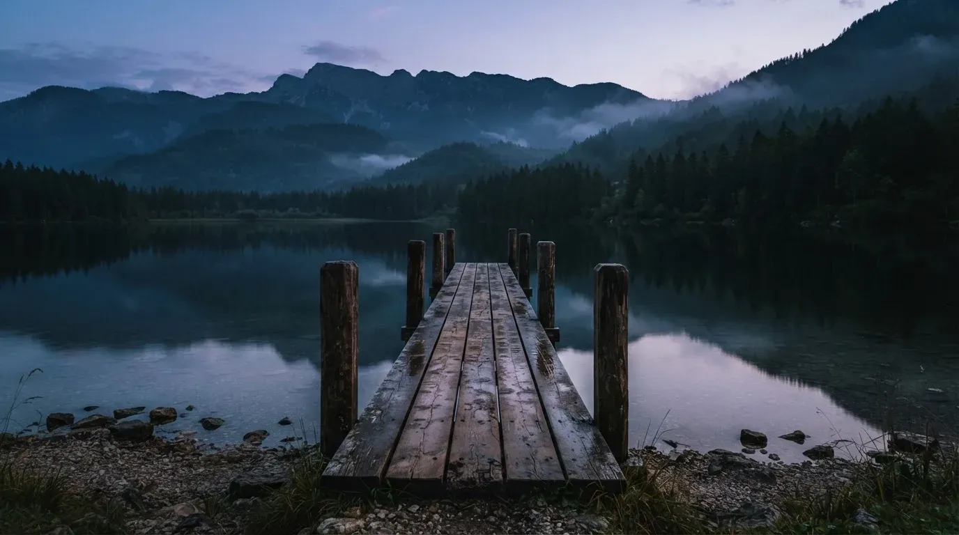 Ein Bergsee in der Dämmerung mit einem leeren Holzsteg, die Wasseroberfläche ist ruhig und Nebel zieht über die Hänge.