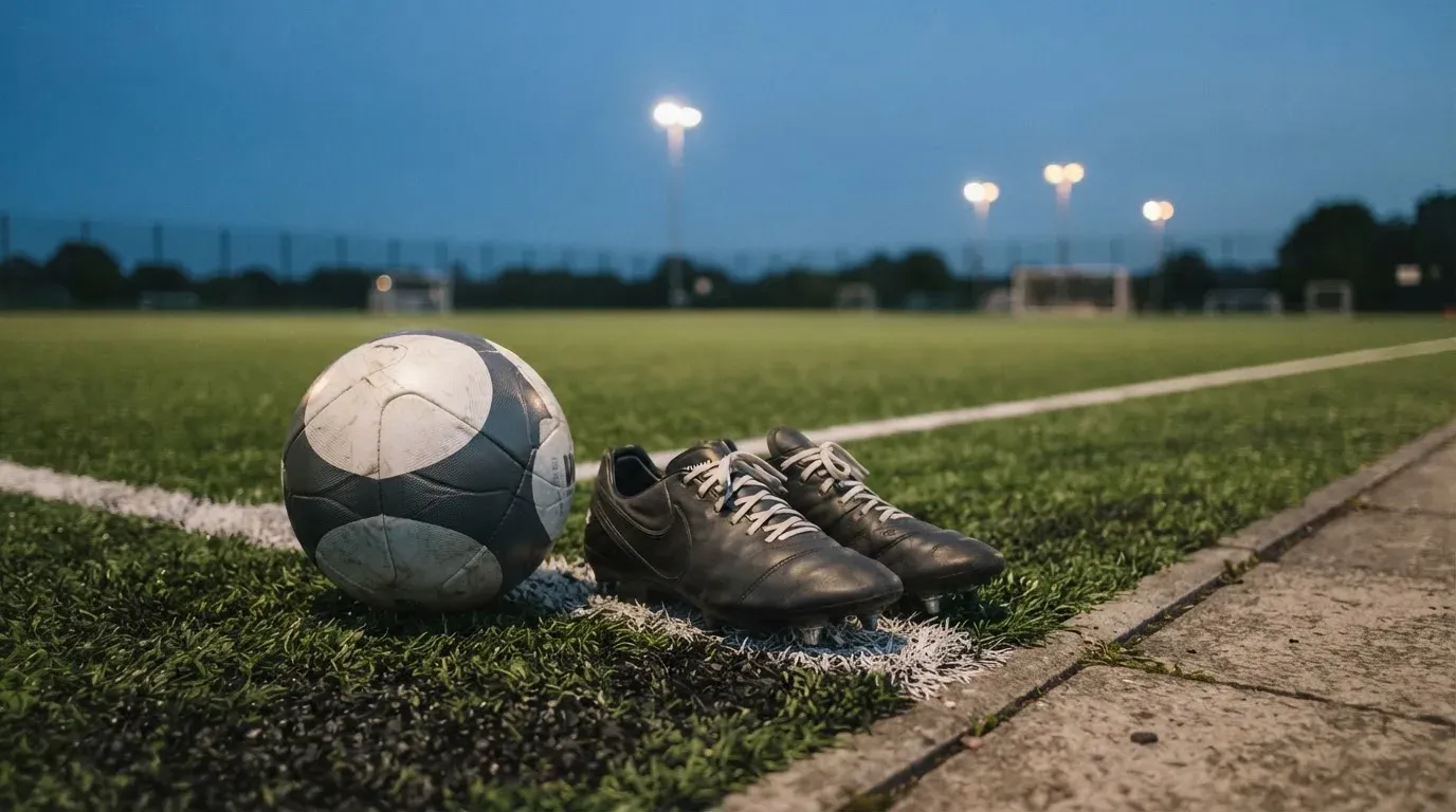 Ein Fussball und ein Paar Stollenschuhe stehen am Rand eines Kunstrasenplatzes, Flutlichtmasten ragen in den Abendhimmel.