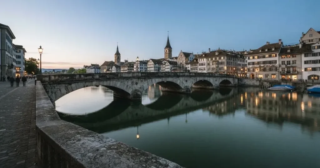 Eine alte Steinbrücke führt über einen Fluss in einer Schweizer Stadt, das Wasser ist ruhig und das Licht kühl.
