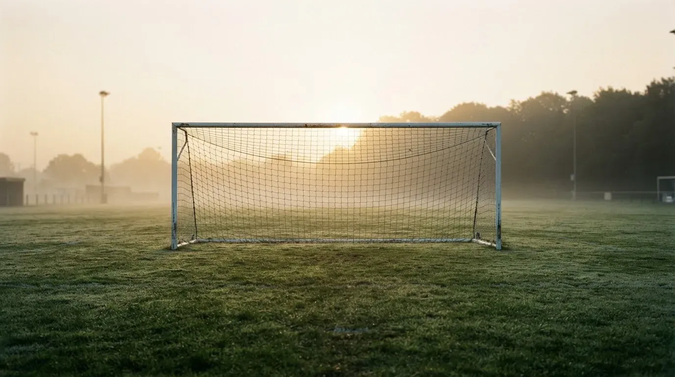 Ein Torpfosten mit Netz steht im Gegenlicht auf einem leeren Fussballplatz, über dem Rasen liegt Nebel.