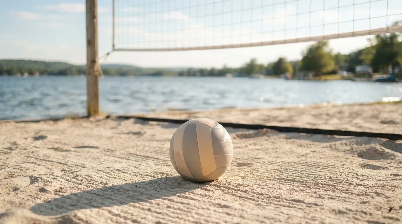 Ein Volleyball liegt auf dem Sand eines Strandfeldes am Seeufer, im Hintergrund glitzert das Wasser.