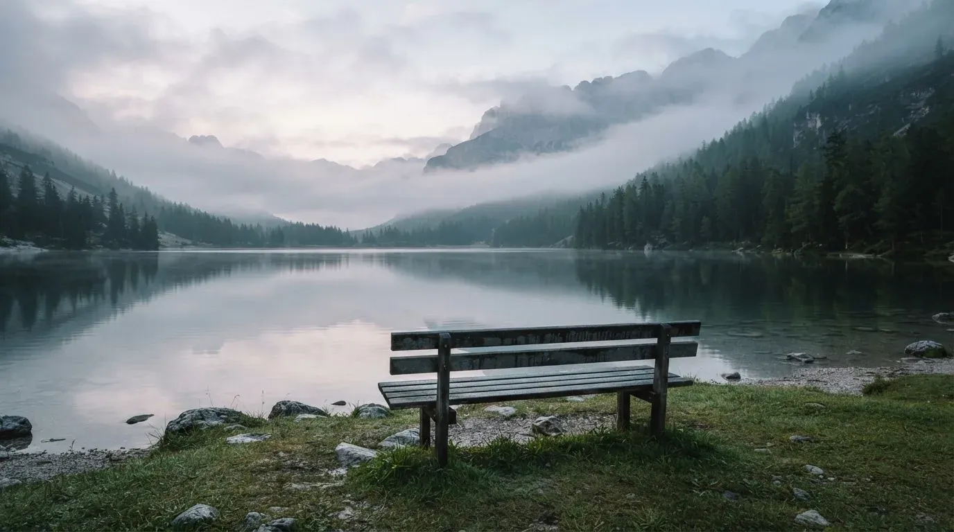 Eine Holzbank am Ufer eines ruhigen Bergsees, am gegenüberliegenden Ufer steigen dunkle Hänge in den Frühnebel