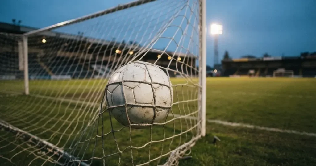 Ein Fussball liegt im Netz eines Tores, Flutlicht und kalte Abendluft liegen über dem leeren Platz