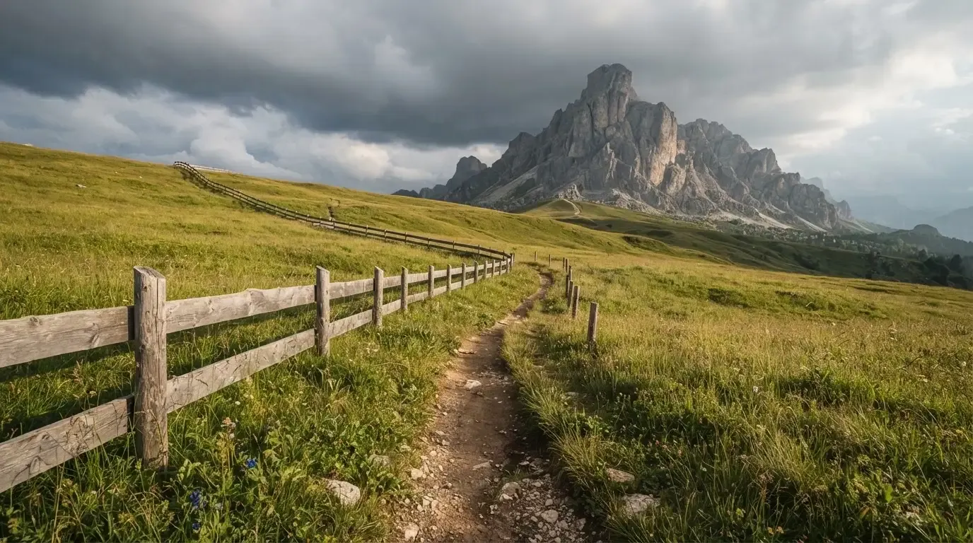 Ein Feldweg führt durch eine Alpwiese mit Holzzaun, im Hintergrund hängen Wolken über einem felsigen Gipfel