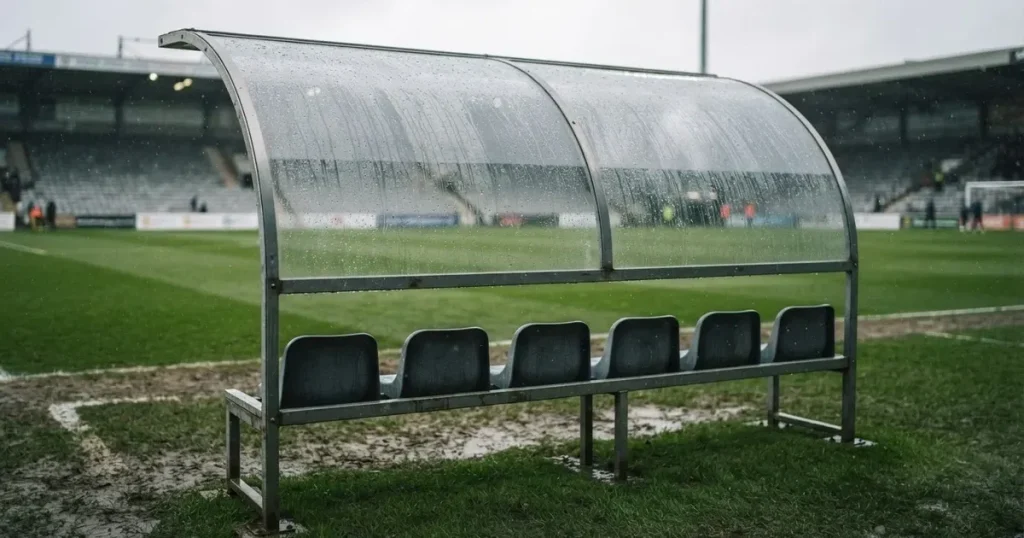Eine leere Ersatzbank unter einem Plexiglasdach am Spielfeldrand, Regentropfen auf der Scheibe, dahinter ein Rasenplatz
