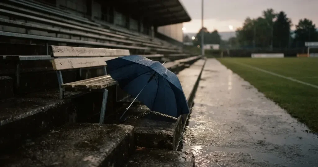 Ein offener Regenschirm lehnt an einer leeren Tribünenbank, Regen fällt auf die Stufen eines Stadions