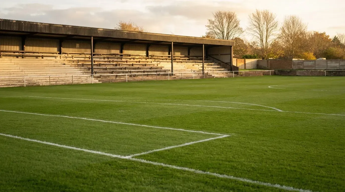 Leere Zuschauerränge eines kleinen Stadions im Herbstlicht, davor ein Rasenplatz mit frisch gezogenen Linien.