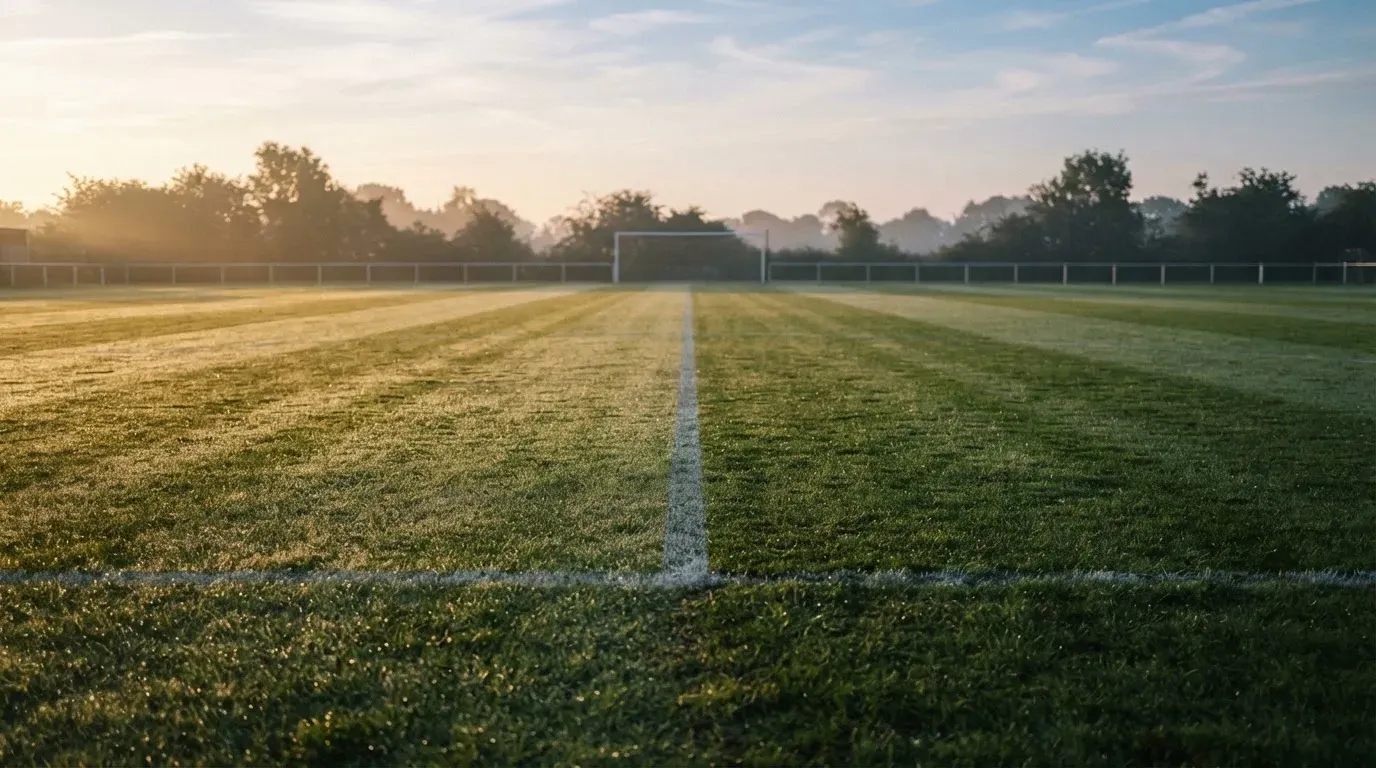 Ein Rasenspielfeld bei Sonnenaufgang, die Mittellinie verläuft quer durchs Bild und Tau glitzert auf dem Gras.
