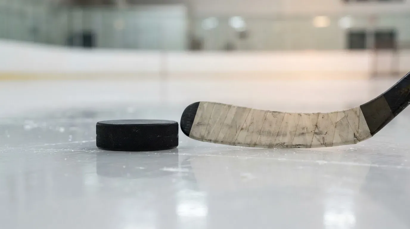 Ein Eishockeypuck und ein Schläger liegen auf einer glänzenden Eisfläche, im Hintergrund verschwimmen leere Banden.