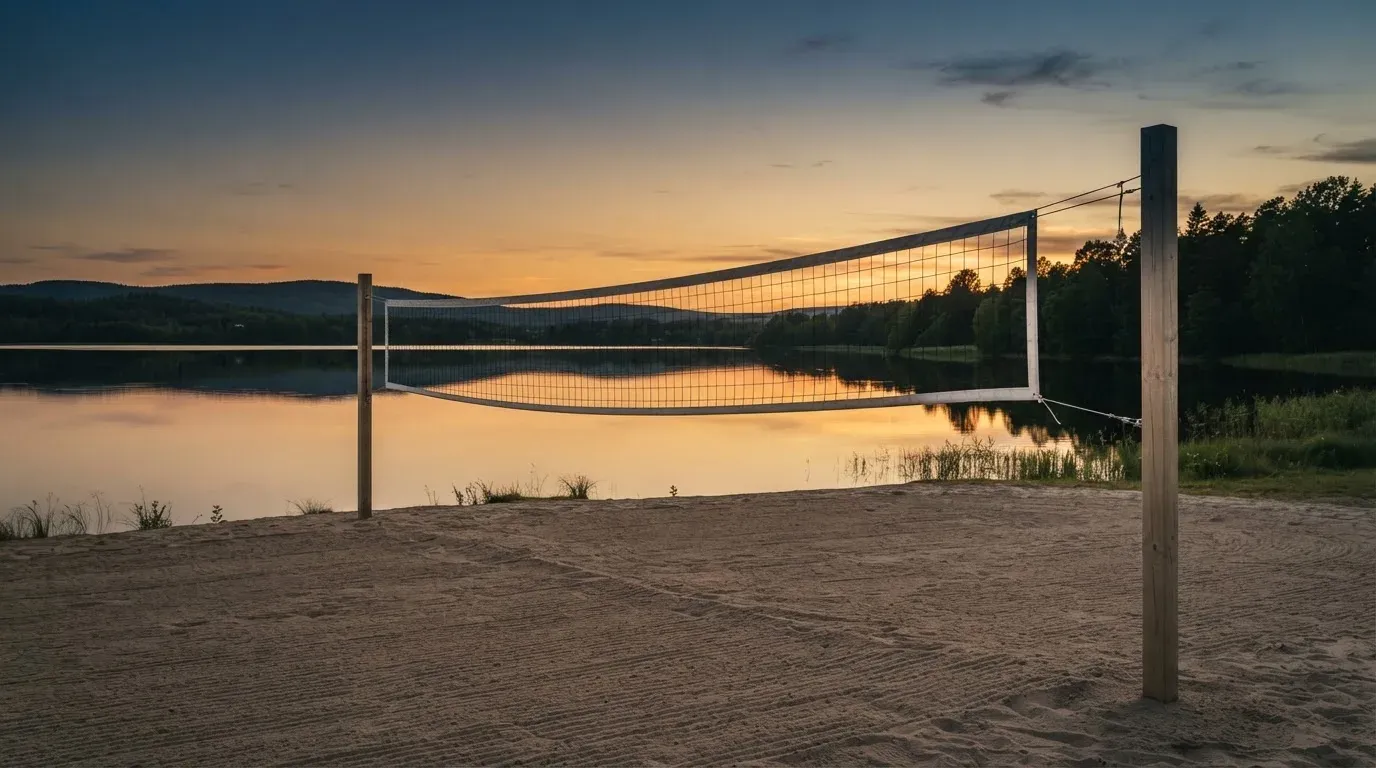 Ein Volleyballnetz steht an einem Seeufer in der Abenddämmerung, der Sand ist leer und das Wasser glatt.