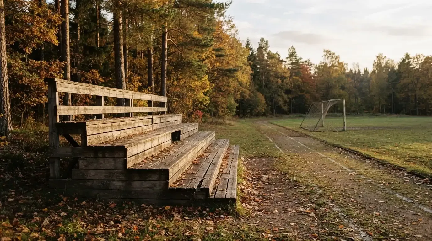 Eine leere Holztribüne steht am Waldrand neben einem kleinen Sportplatz im Herbstlicht.
