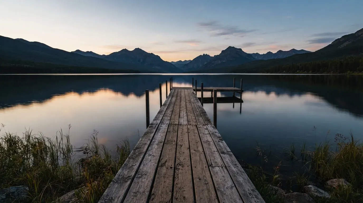 Ein leerer Holzsteg ragt am Abend in einen ruhigen Bergsee, das Wasser spiegelt den Himmel.