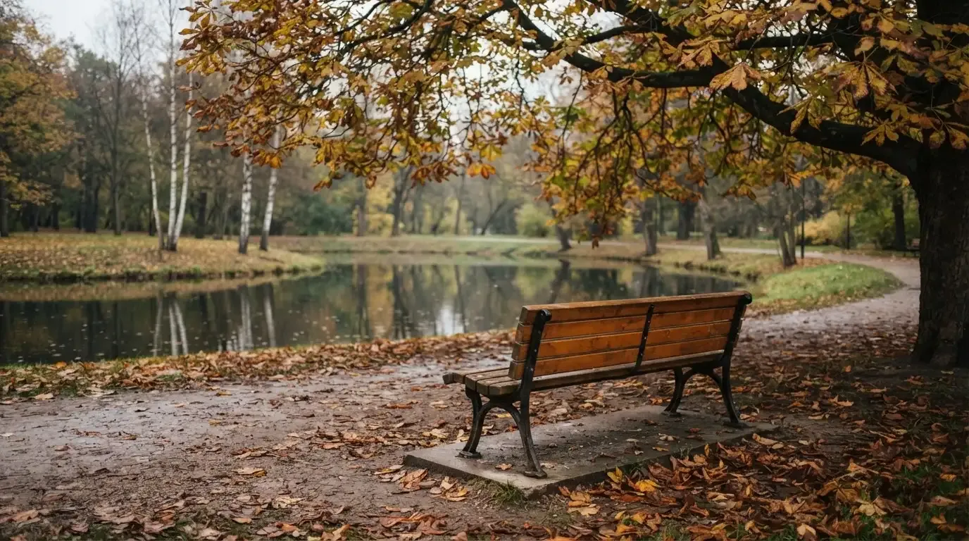 Eine Parkbank unter einer Kastanie, Herbstlaub bedeckt den Kiesweg, im Hintergrund ein stiller Teich.