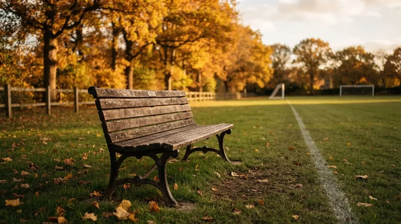 Eine hölzerne Bank steht an der Seitenlinie eines leeren Sportplatzes, dahinter Bäume im Herbstlicht.